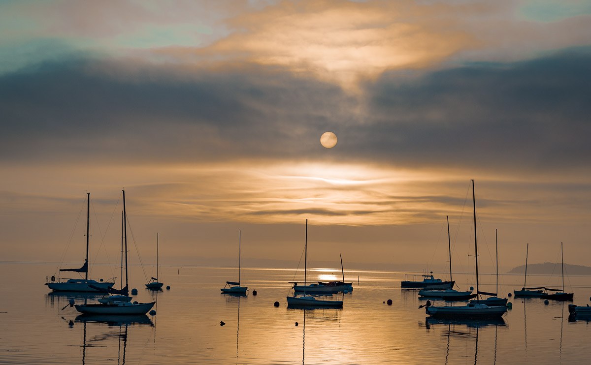 disk of sun filtered through morning clouds with sailboats at anchor below