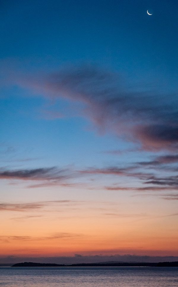 a sliver of a moon rides high above colorful pre-sunrise clouds