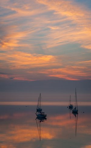 four sailboats at anchor on smooth water under colorful dawn clouds